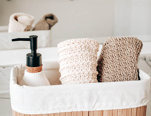 A picture of two crochet towels in a basket with a soap container on top of a bathroom counter with a mirror in the background.