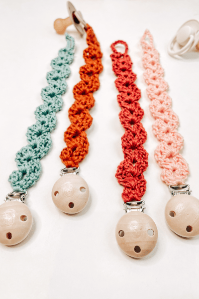 An image that shows 4 crochet pacifier clips in light blue, orange, terracotta and light pink.