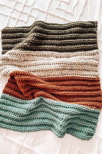 An image of a crochet baby blanket with colour stripes. Green, off white, orange and baby blue on a white bed.